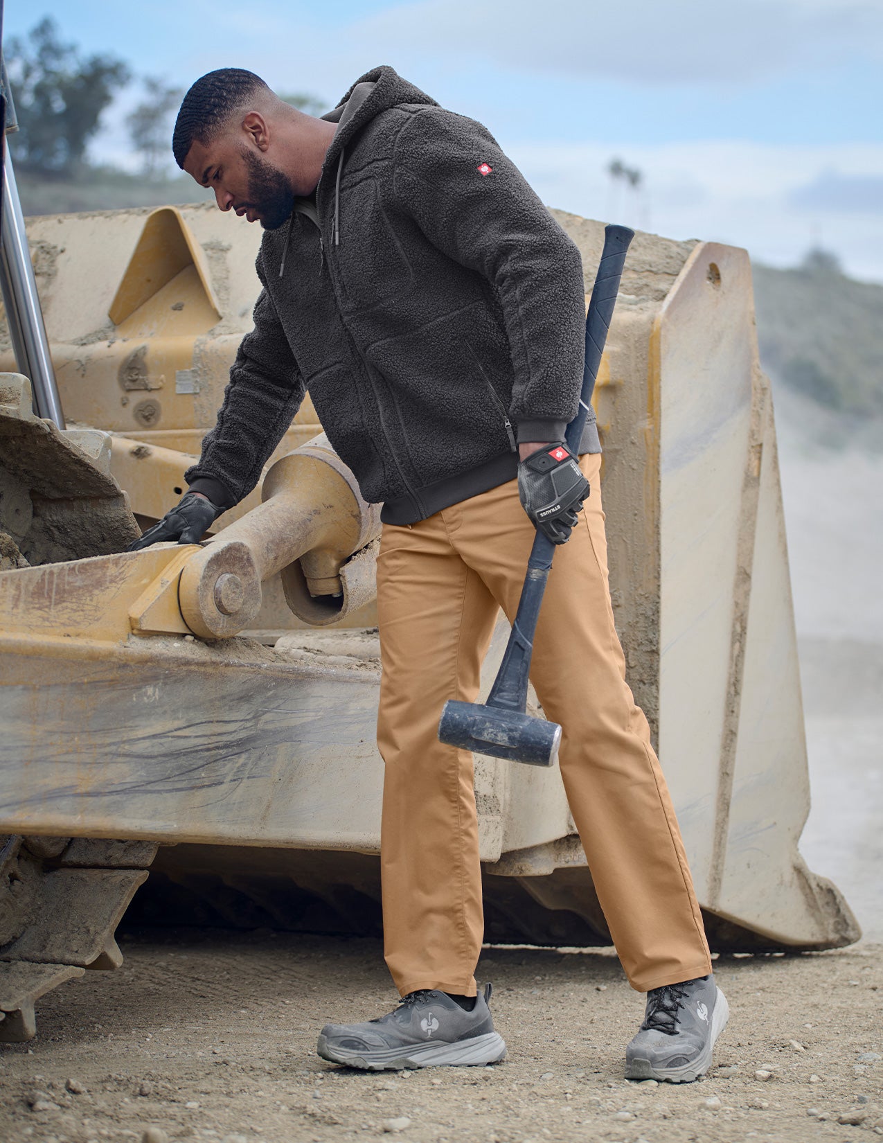 Person in work attire with a tool, standing next to heavy machinery.