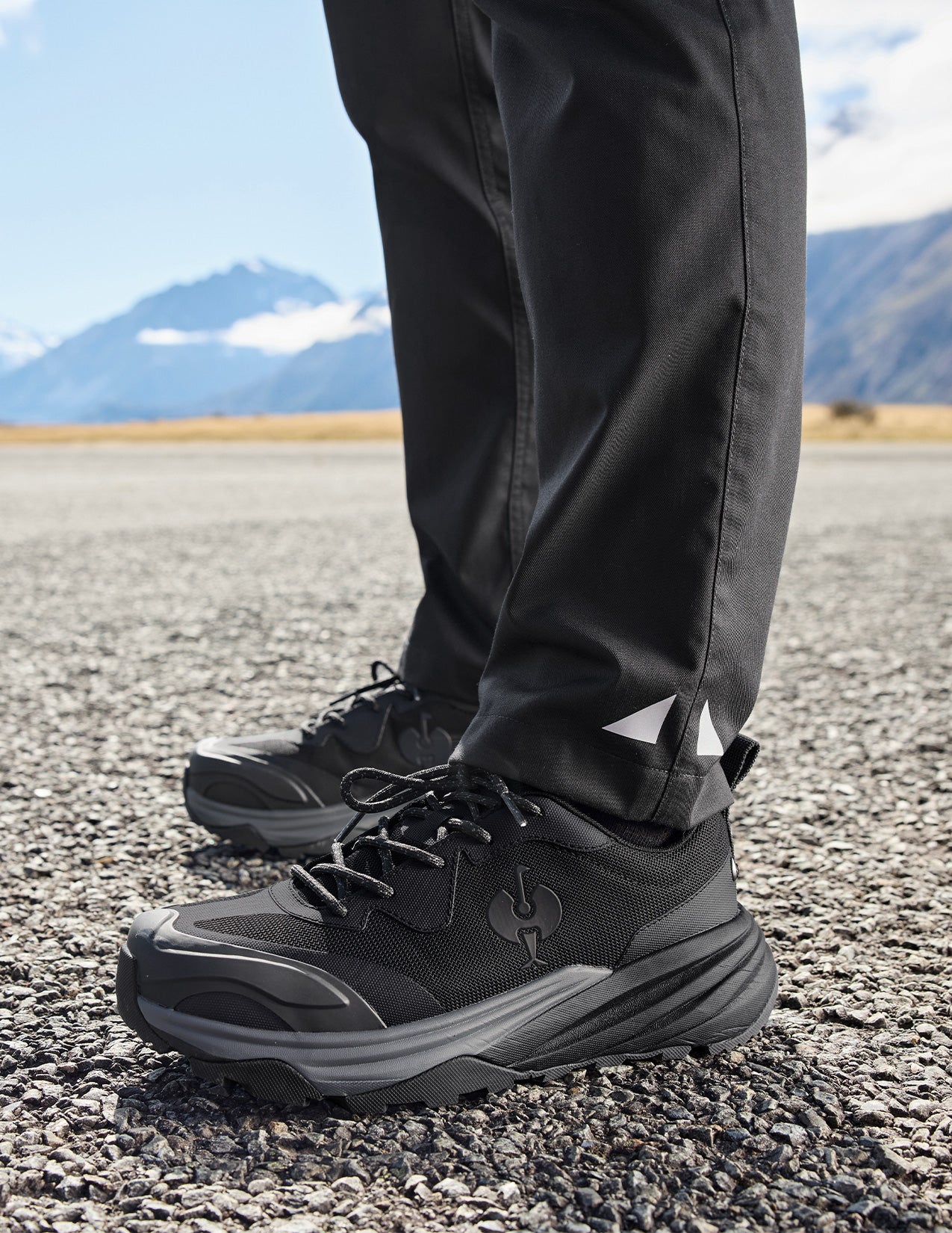 Black hiking shoes and pants on a person standing on a gravel surface with mountains in the background