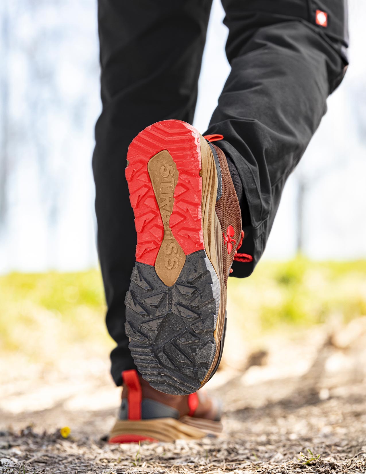 Close-up of a person wearing desertbrown shoes with red soles on a natural path.