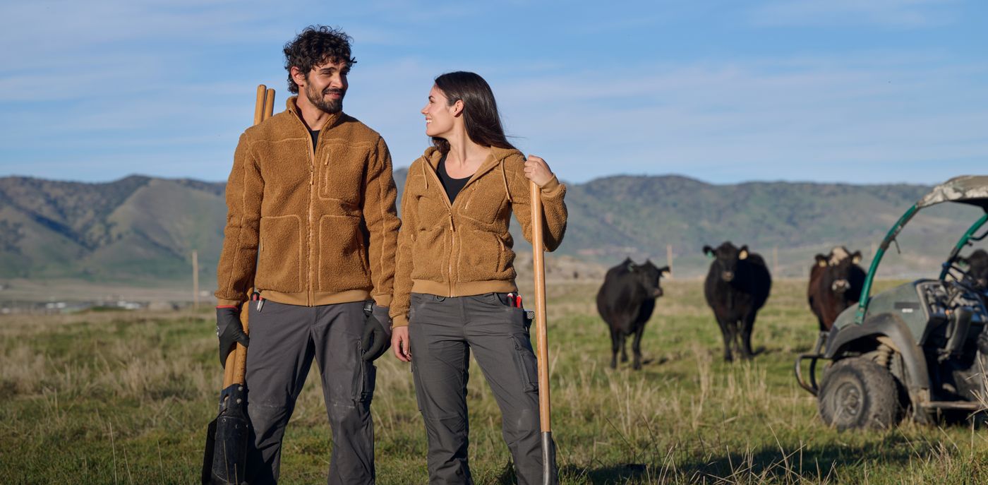 Two people in brown jackets standing in a field with cows and a vehicle in the background.