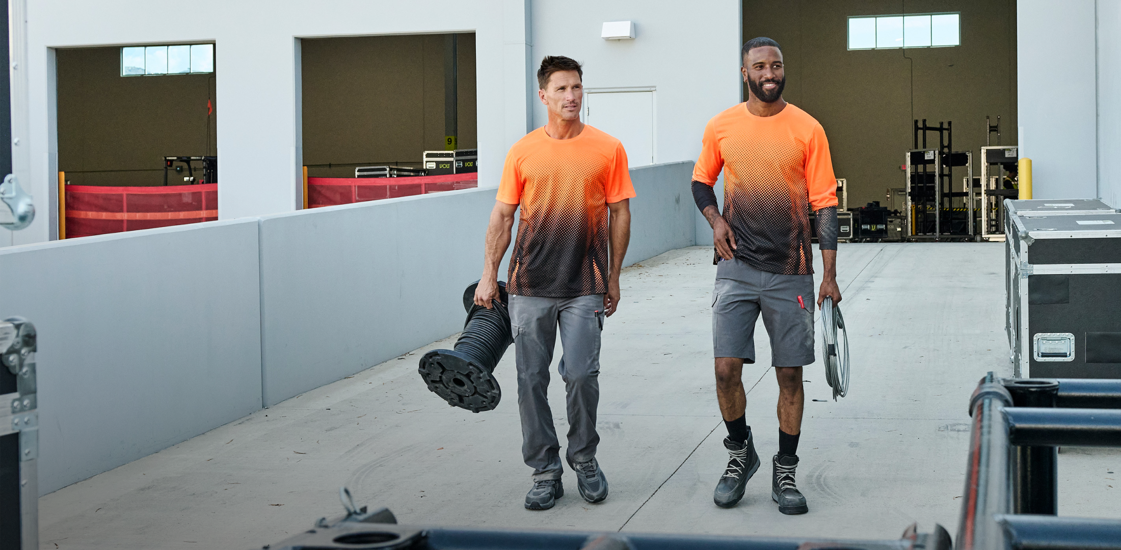 Two men in matching orange and black gradient t-shirts standing in a warehouse.