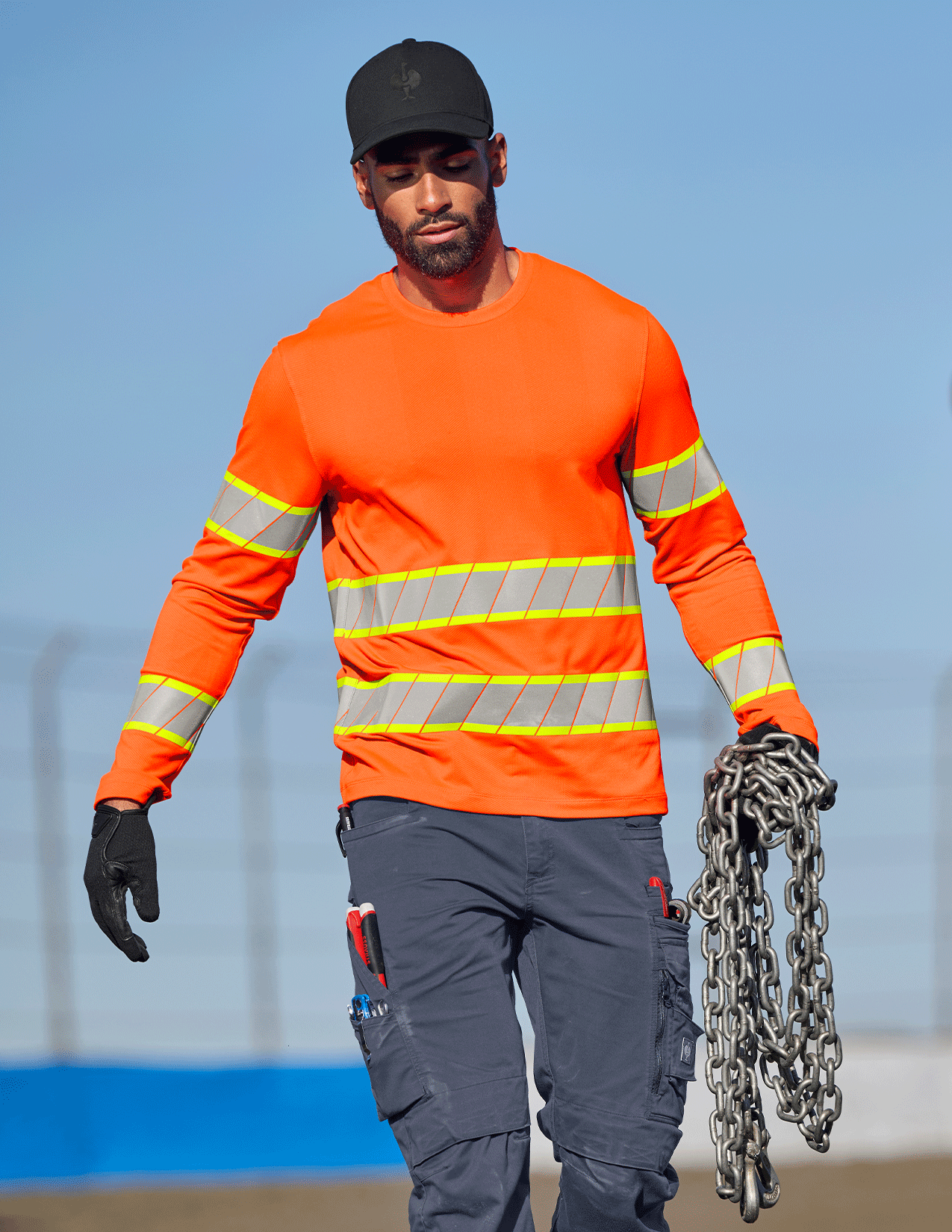 Person wearing an orange safety shirt with reflective stripes, holding chains against a clear blue sky.