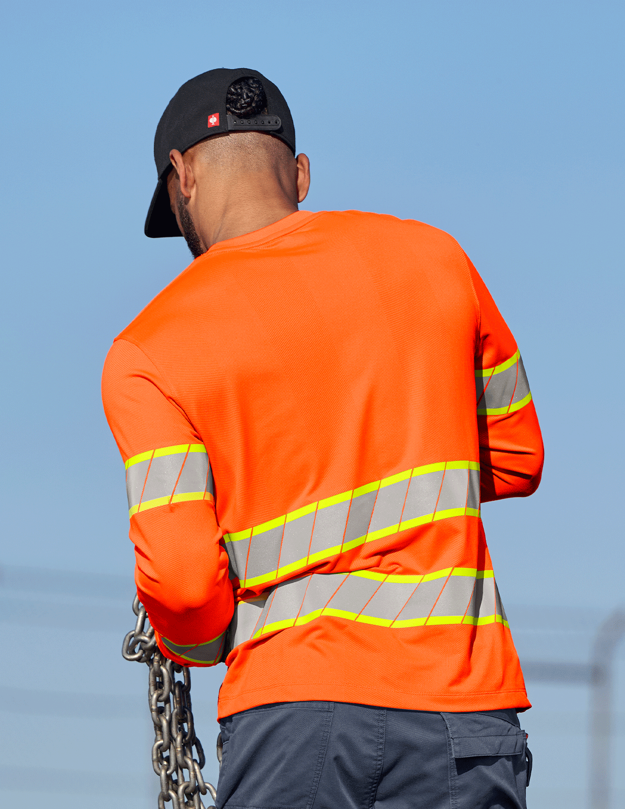 Person wearing an orange safety jacket with reflective stripes against a clear blue sky.