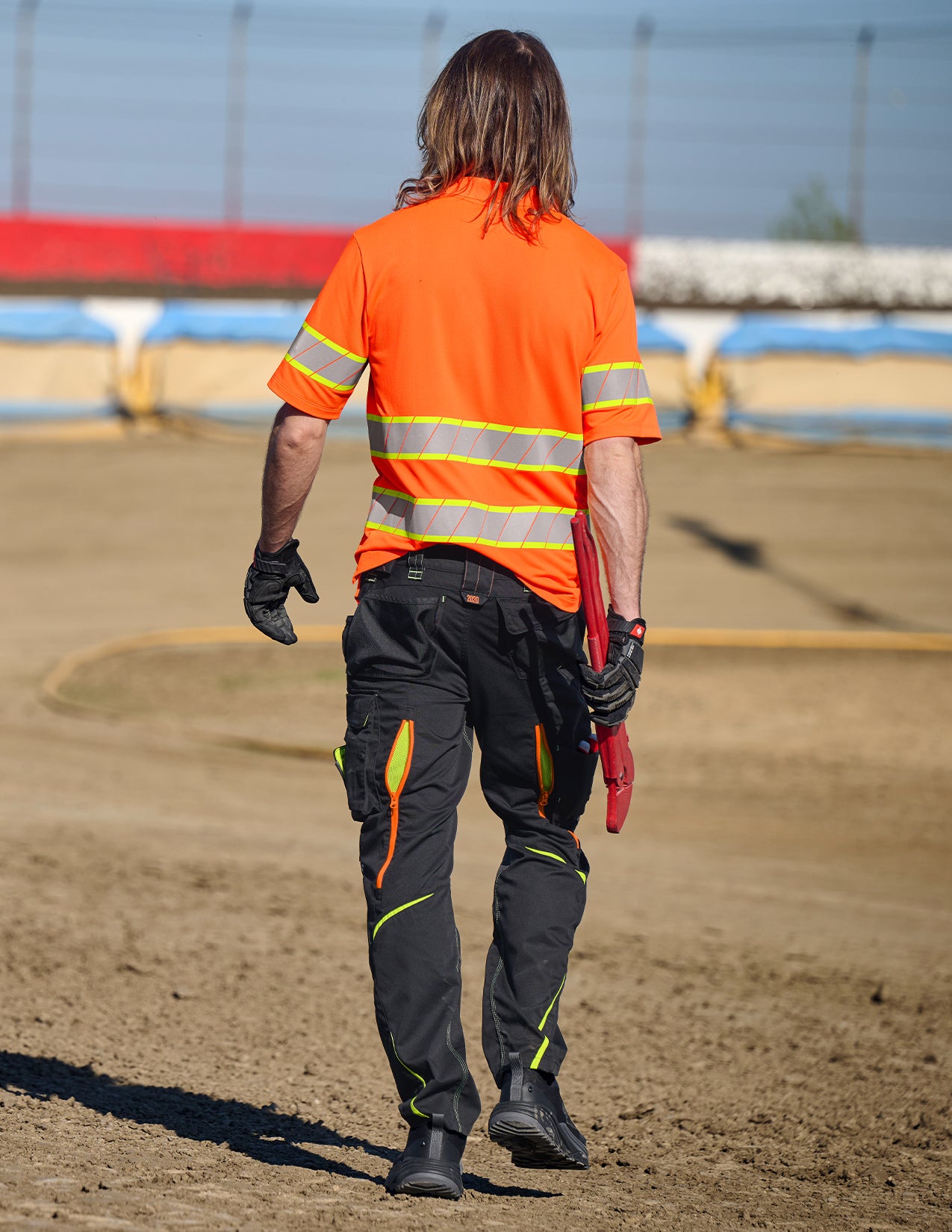Person wearing an orange safety shirt and black pants with reflective stripes, holding a red tool on a dirt track.