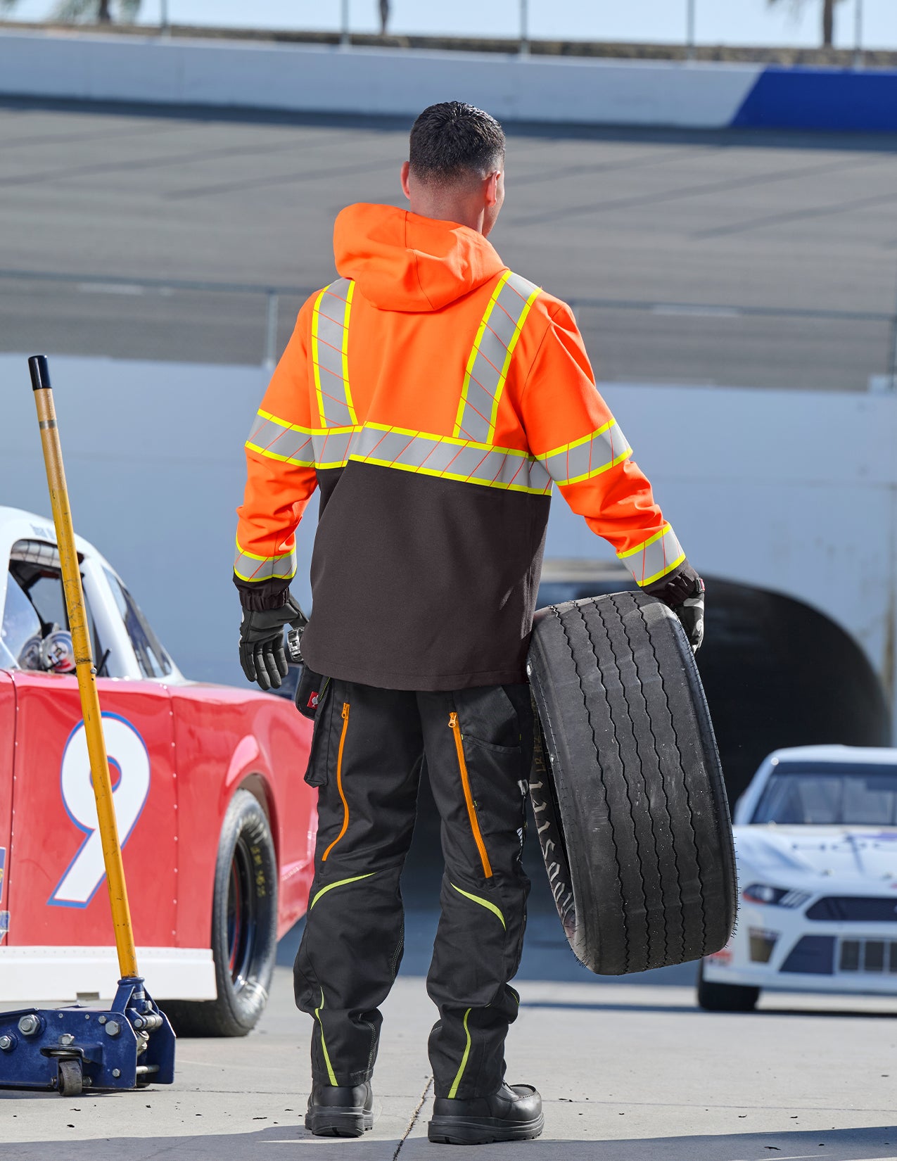 Person in safety gear holding a tire on a race track with cars in the background