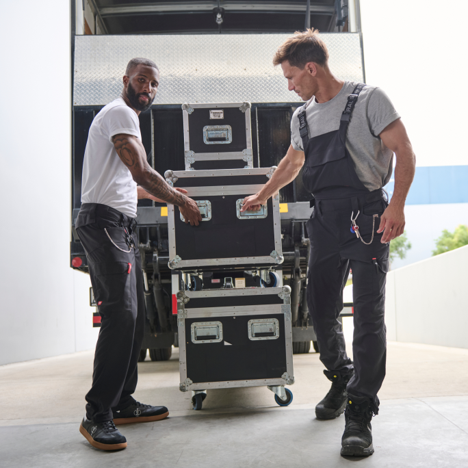 Two men moving a stack of black flight cases in front of a bus.