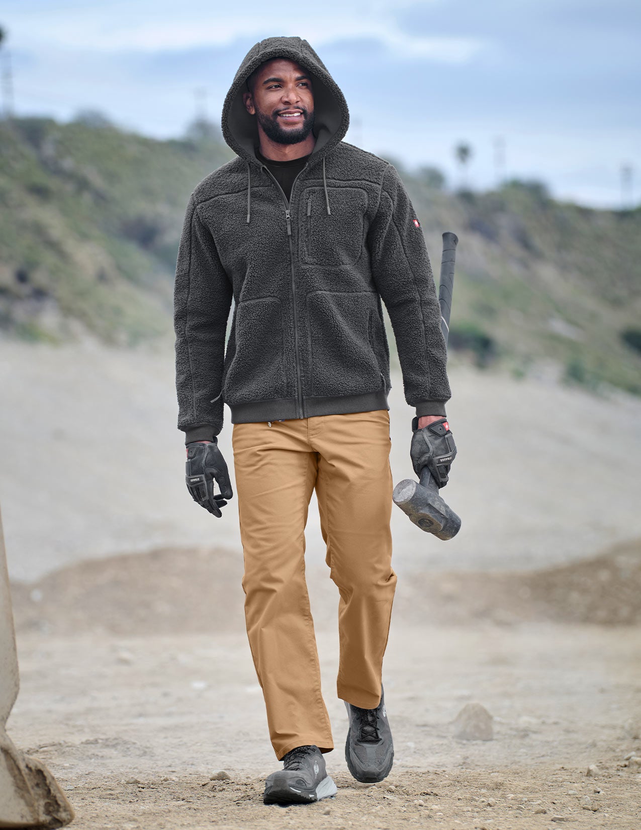 Man wearing a dark gray jacket, tan pants, and gloves standing outdoors with a blurred background