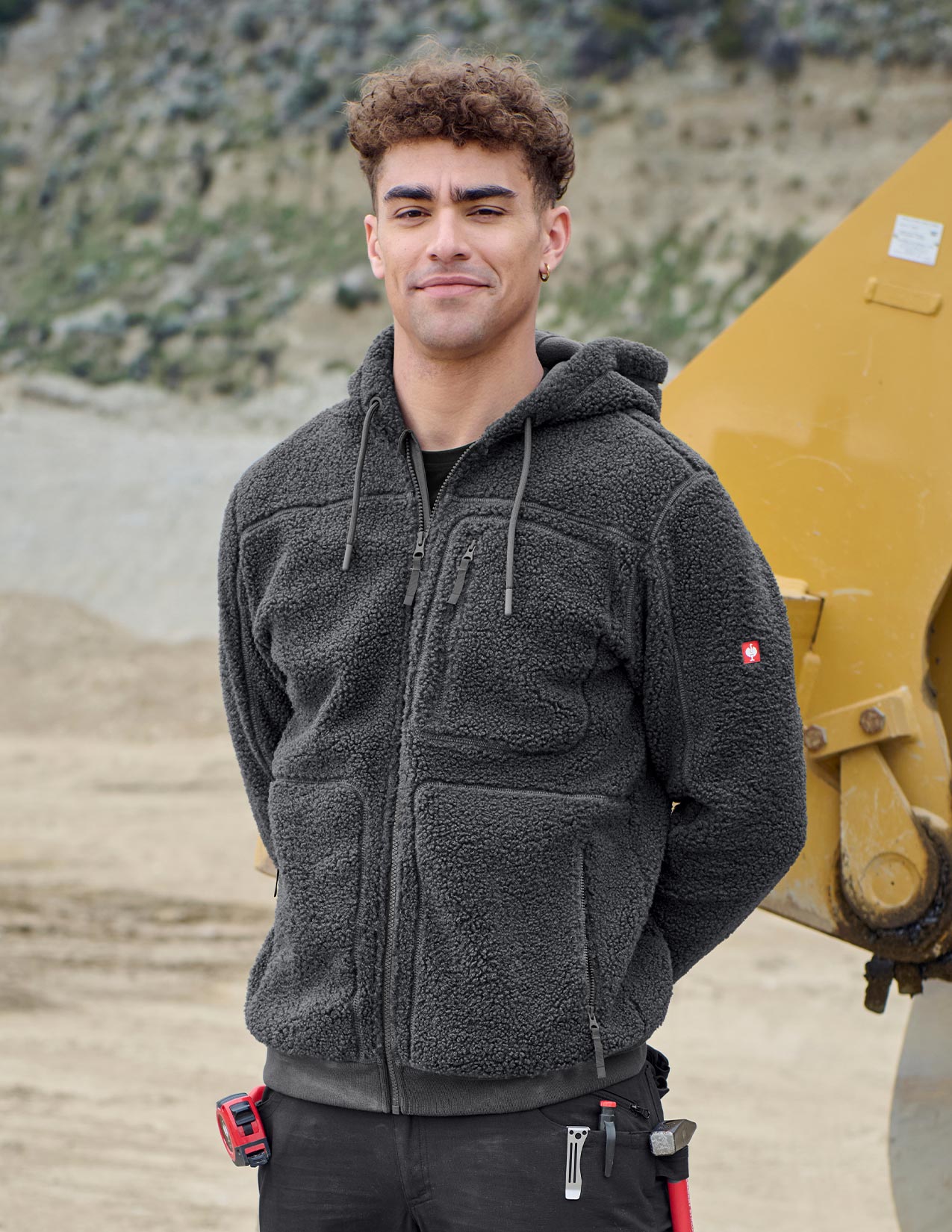 Person wearing a dark gray jacket, standing in front of a yellow piece of machinery.