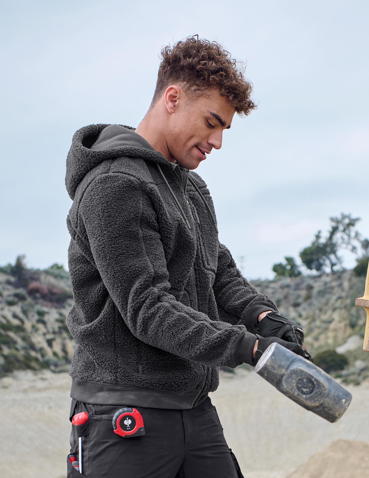 Person wearing a dark gray jacket holding a hammer outdoors.