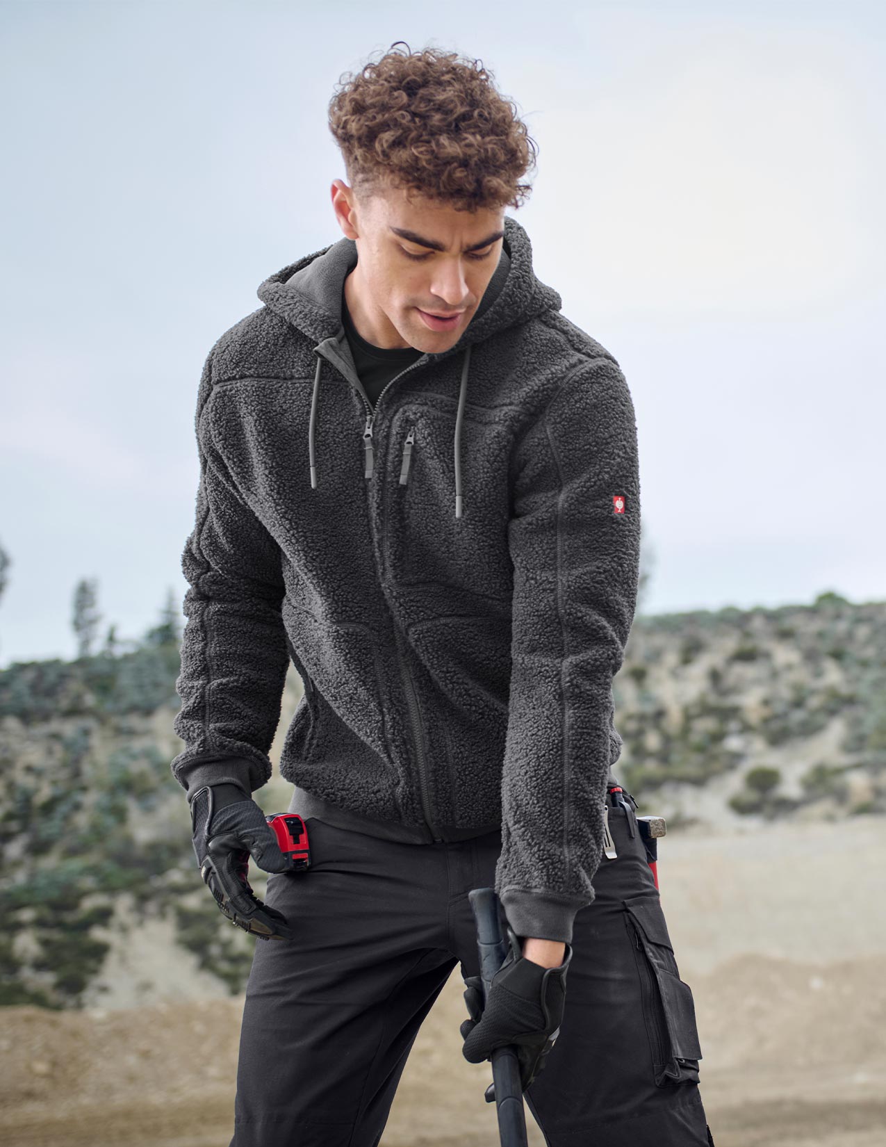 Man wearing a dark gray jacket and black pants with tools, standing outdoors.