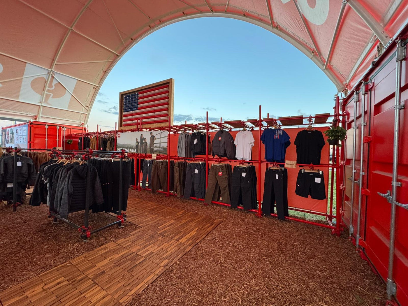 Outdoor clothing display with various jackets and shirts under a red tent.