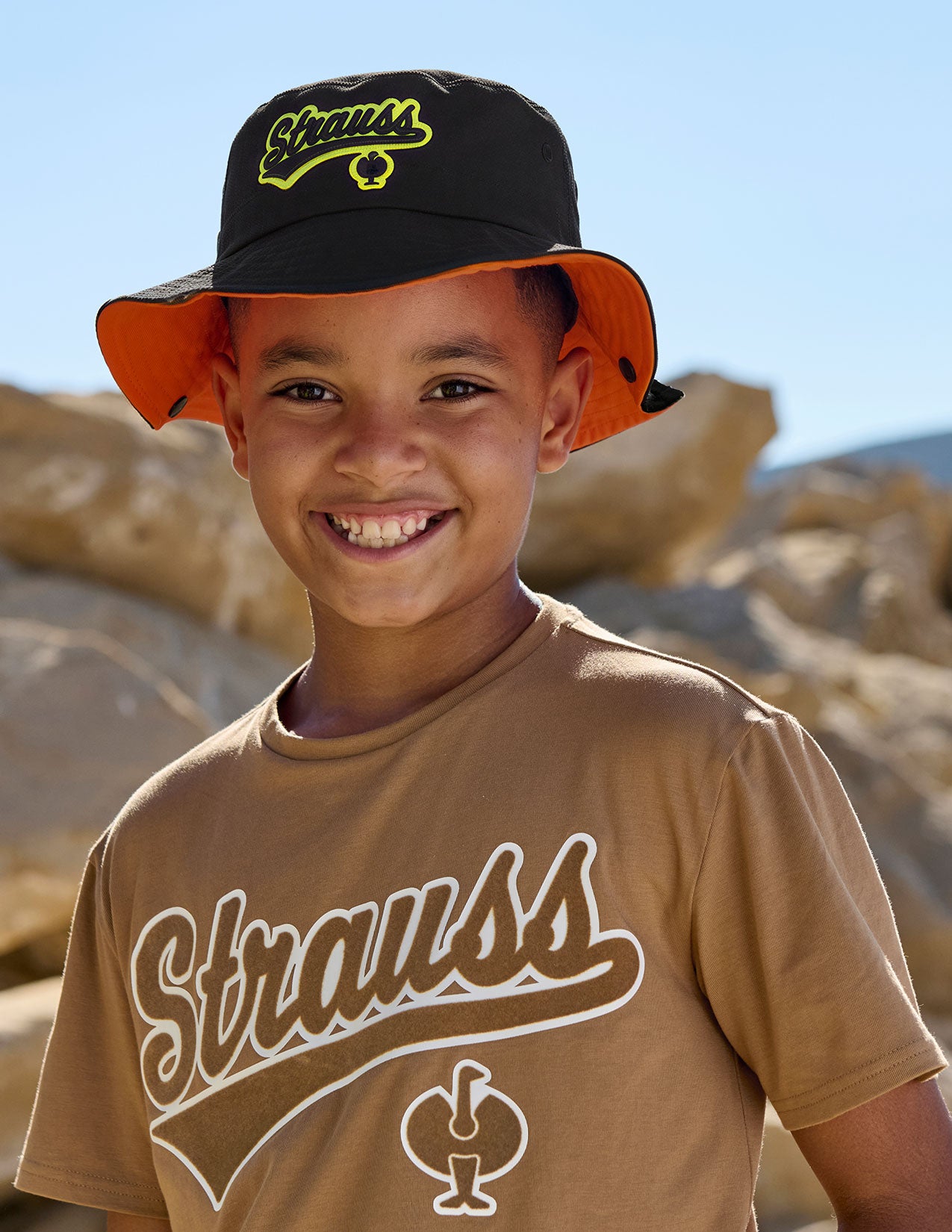 Child wearing a black and orange hat with 'Strauss' branding, standing in a desert-like setting.
