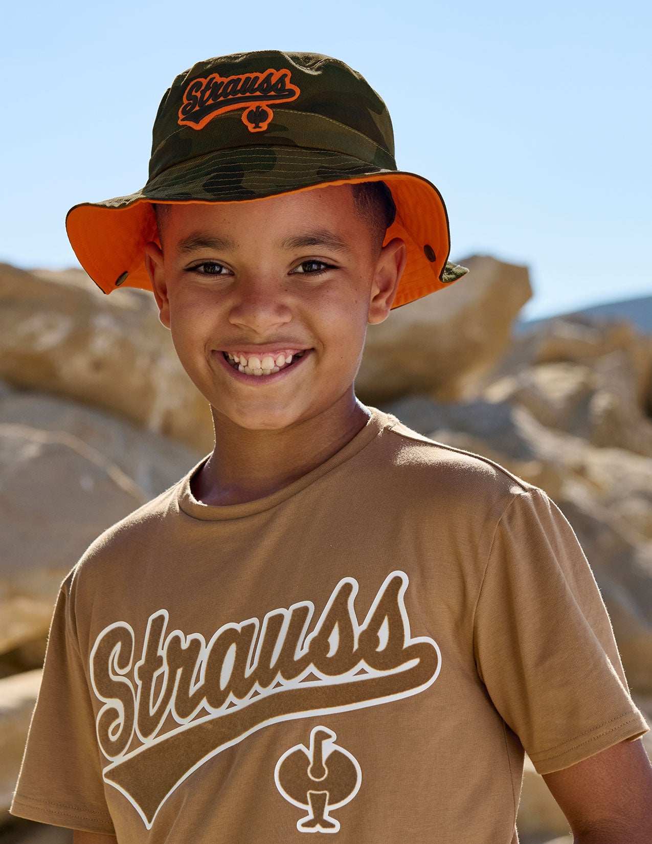 Child wearing a brown 'Strauss' t-shirt and black hat in a desert setting.