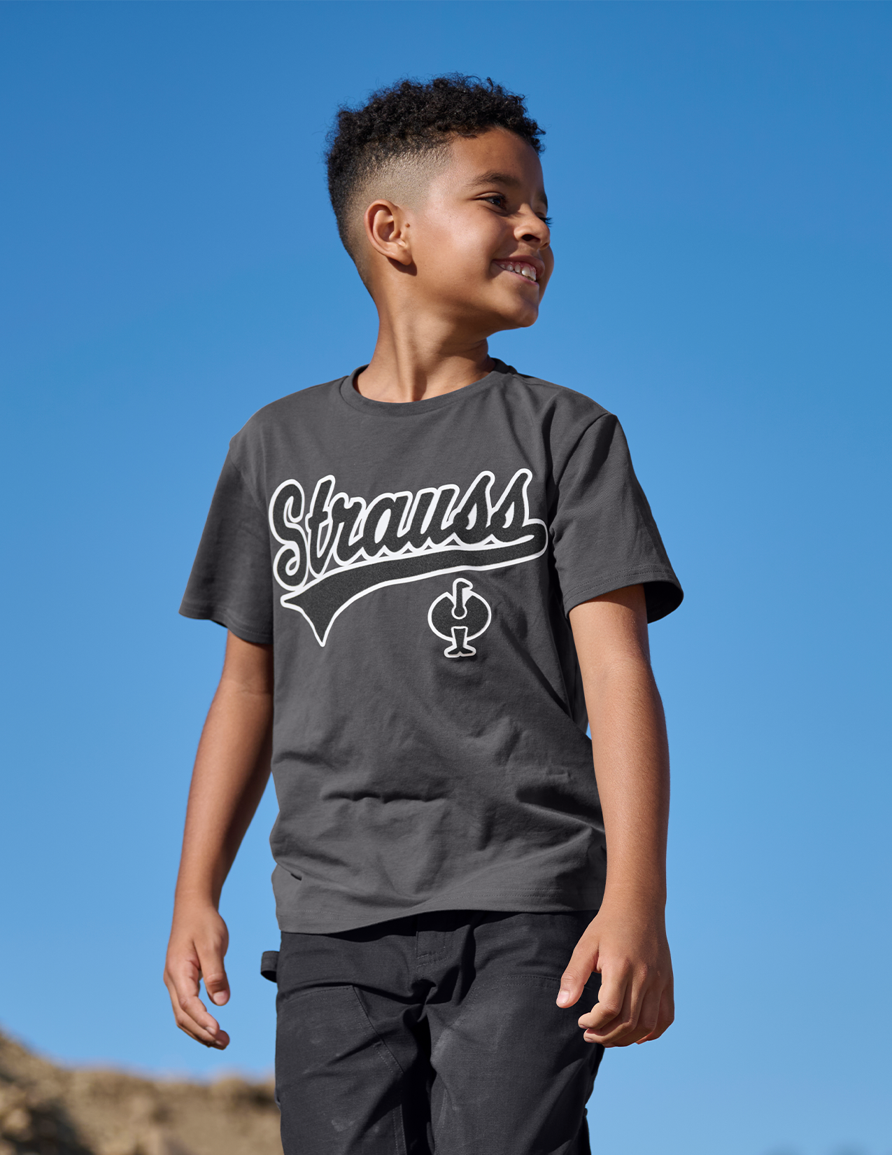Child wearing a dark gray t-shirt with 'Strauss' logo against a clear blue sky