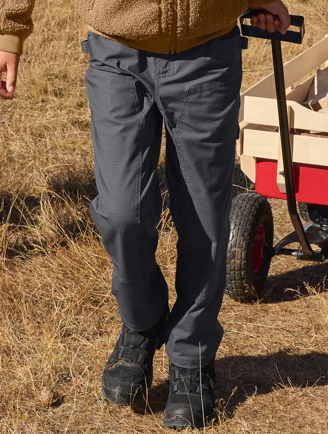 Child wearing grey pants and black shoes in a field with a red wagon in the background.