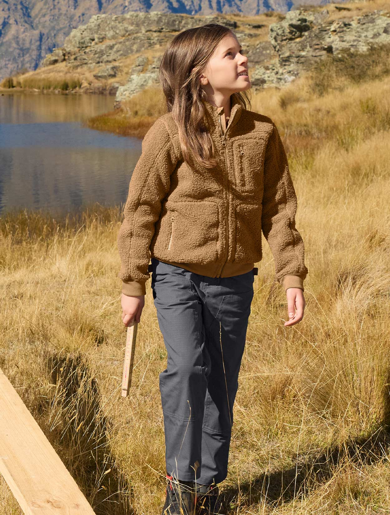 Child wearing a brown jacket and gray pants standing in a natural setting with a lake and mountains in the background.