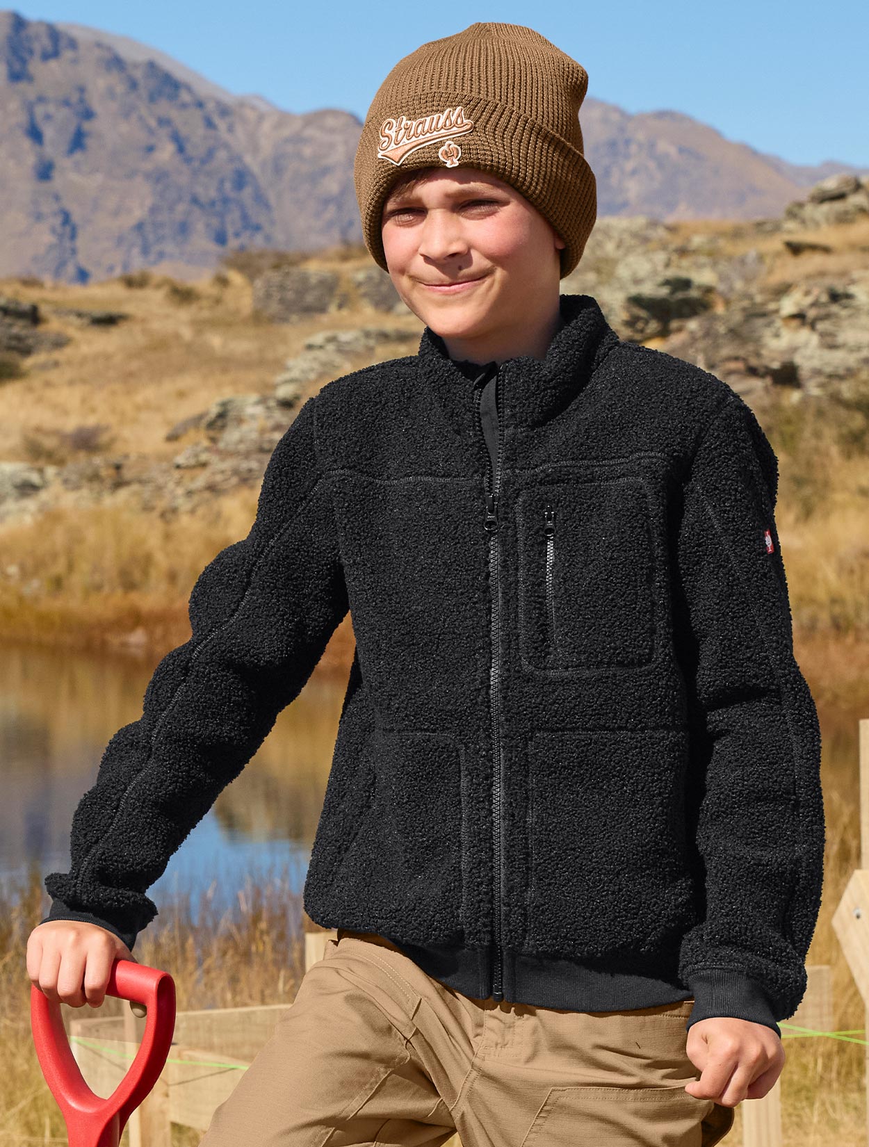 Child wearing a black jacket and brown beanie standing outdoors with mountains in the background