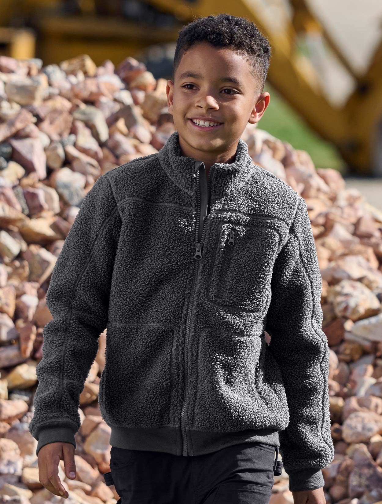 Child wearing a dark gray fleece jacket standing in front of a pile of stones.