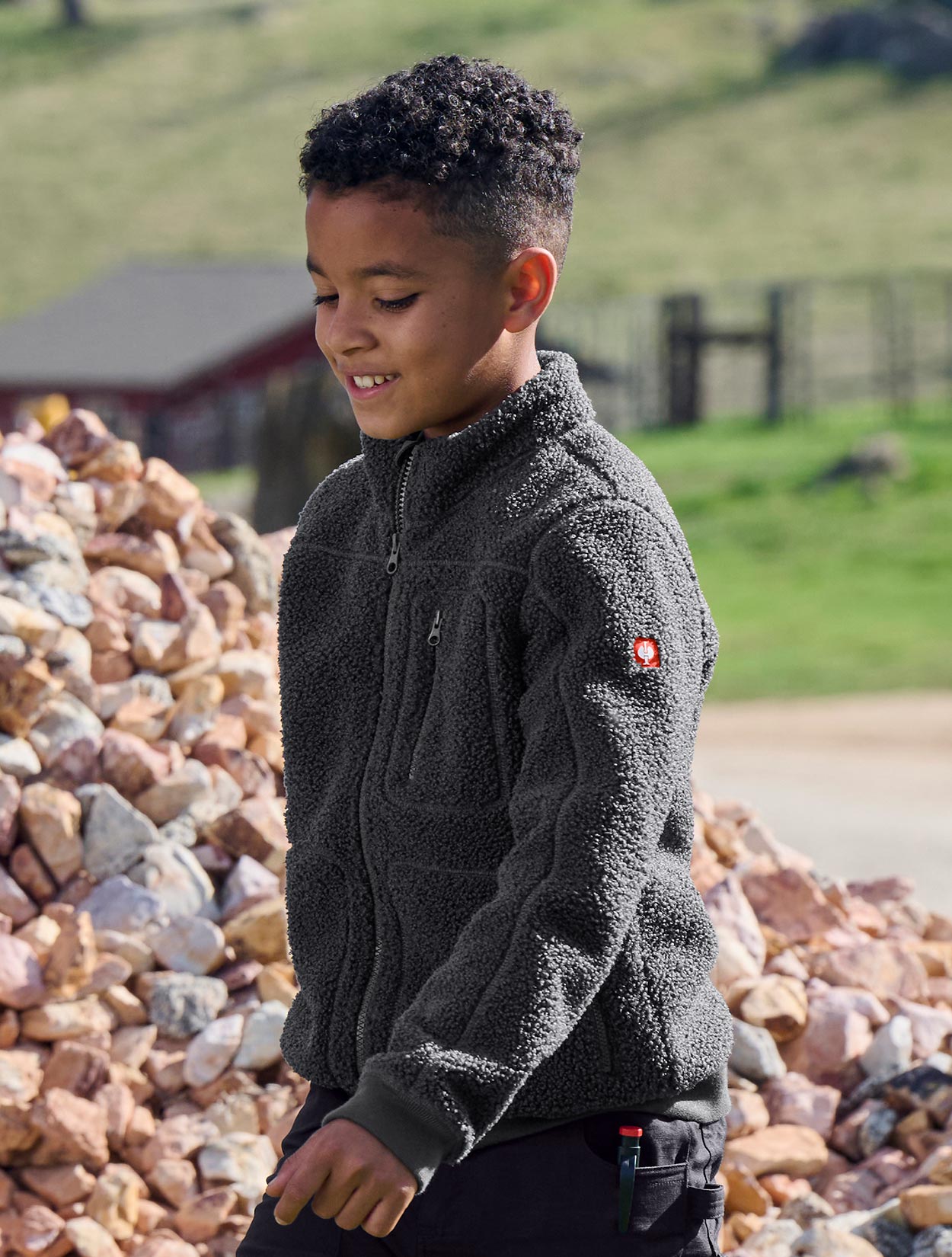 Child wearing a dark gray fleece jacket with a brand logo, standing outdoors near rocks and grass.