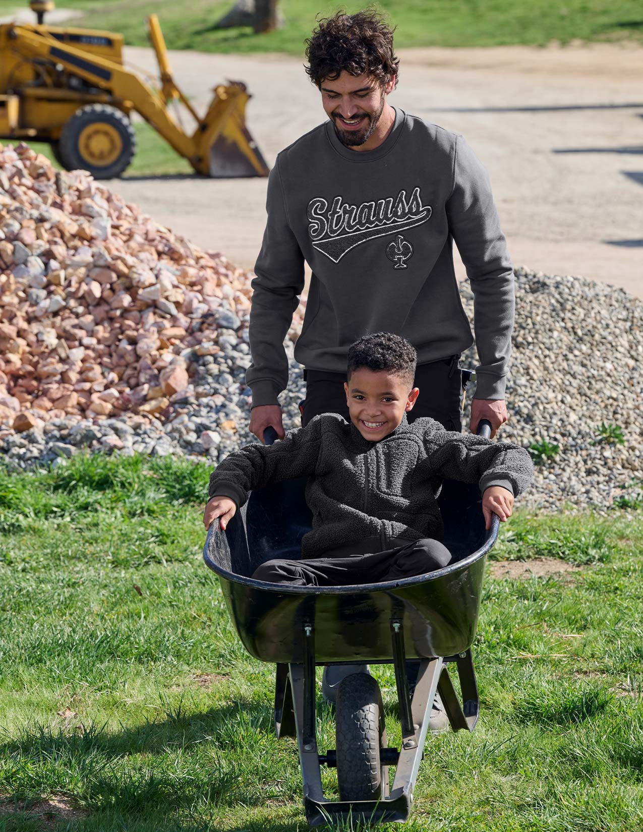 Man pushing a child in a wheelbarrow with construction equipment in the background