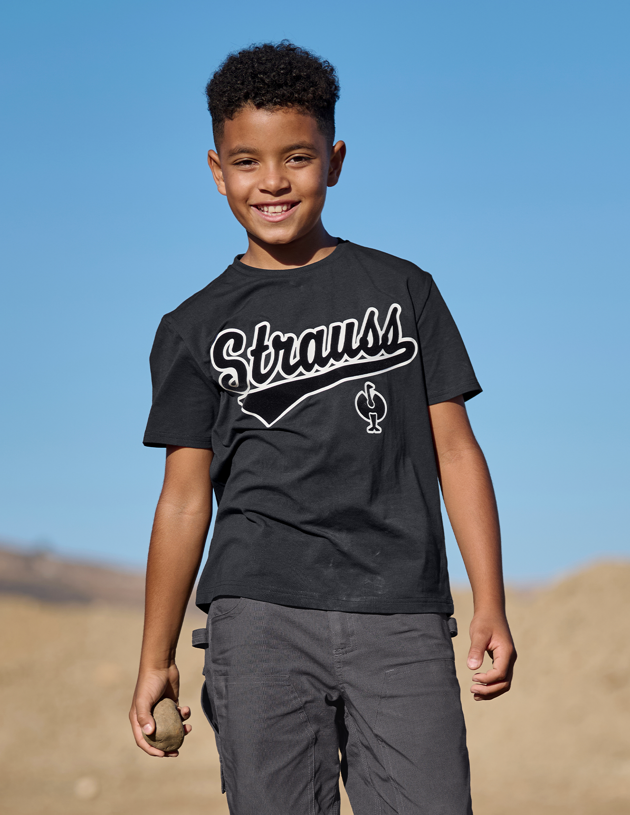 Child wearing a black t-shirt with 'Strauss' logo outdoors against a blue sky.
