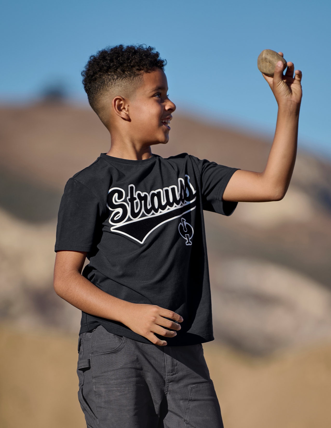 Child wearing a black 'Strauss' t-shirt holding a rock outdoors.