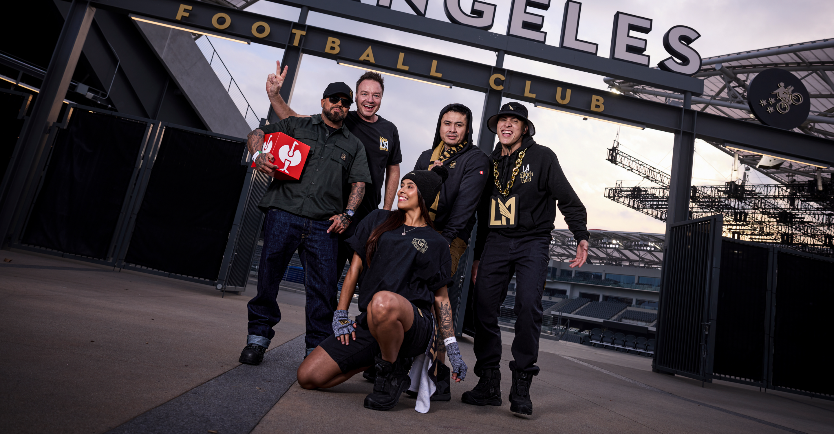 Five people wearing black LAFC attire pose and smile outside a stadium entrance. One person kneels in front, while others stand behind, with two flashing peace signs. The FOOTBALL CLUB sign is visible above them.
