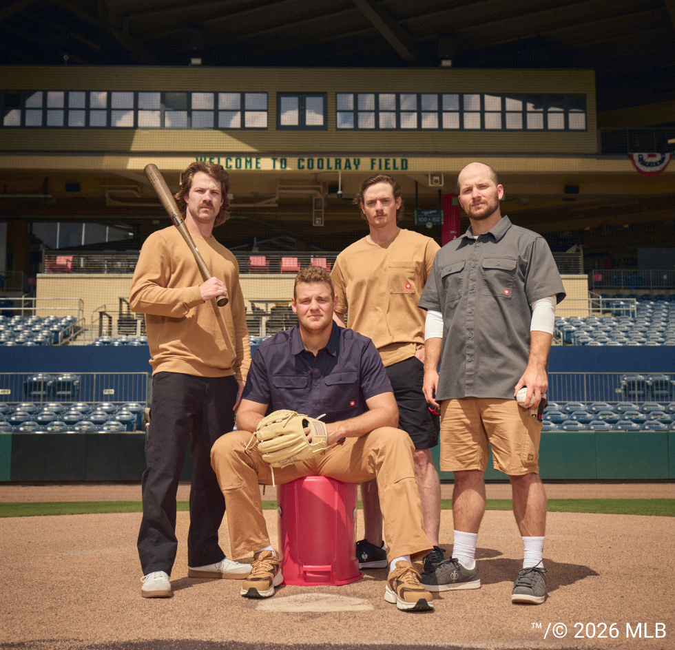 Four men posing in baseball field uniforms in front of a baseball stadium. Two wear tan and black uniforms, one wears a navy and tan uniform sitting on a red stool, and another wears a gray and khaki uniform holding a white baseball.