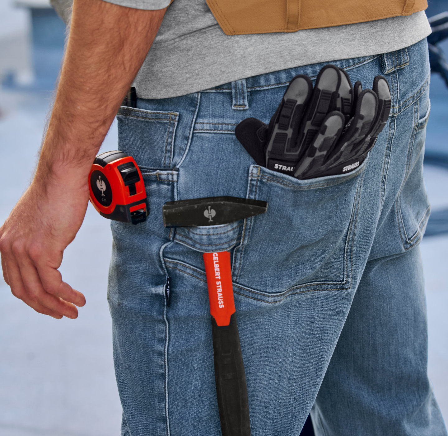 A person wearing jeans with a tape measure, black work gloves, and a hammer tucked into their tool belt, seen from the side and back.