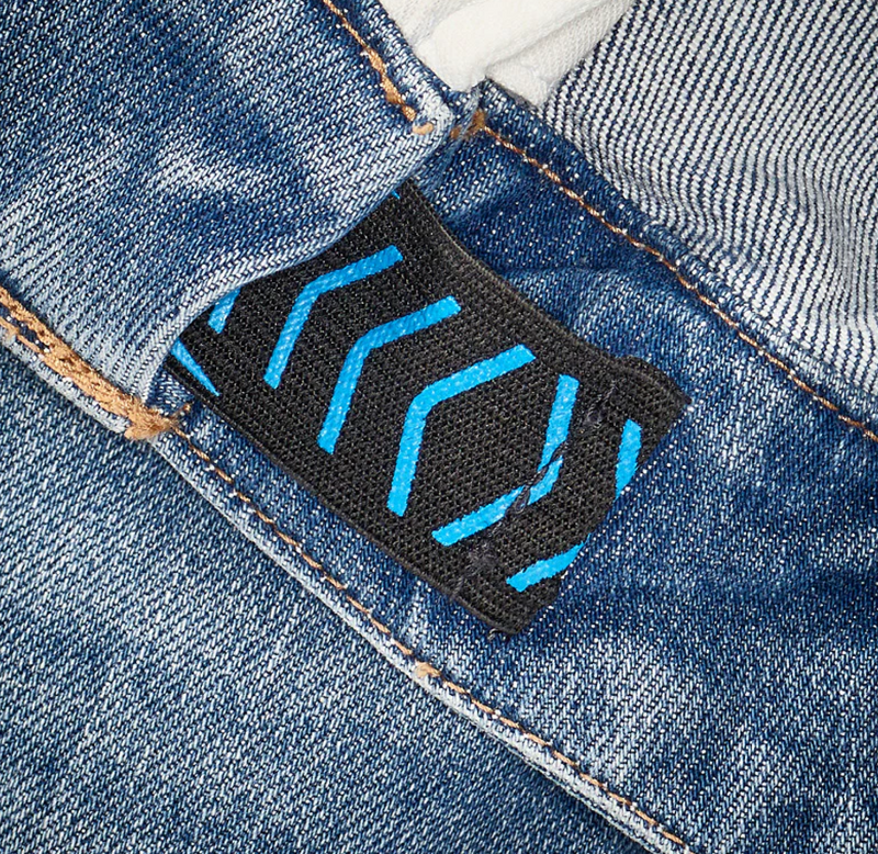 Close-up of blue denim jeans with a black fabric tag featuring blue stitched chevron designs on the waistband. White fabric is partially visible above the jeans.
