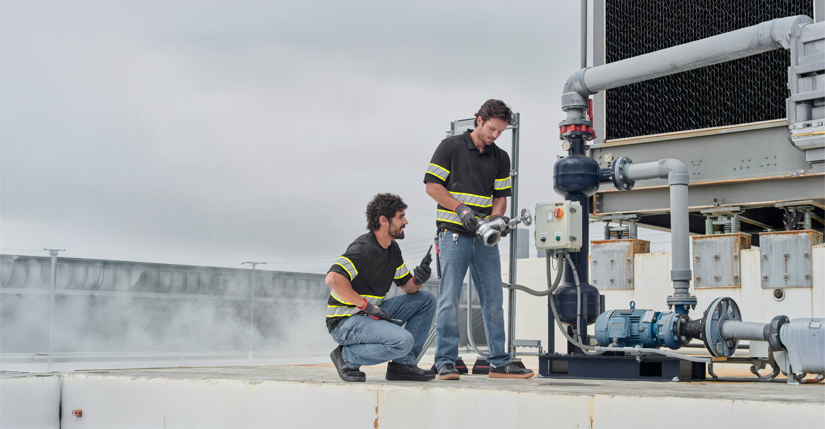 Two workers in reflective shirts inspect and operate industrial equipment on a rooftop, with pipes and machinery visible. Steam rises near them under a cloudy sky.