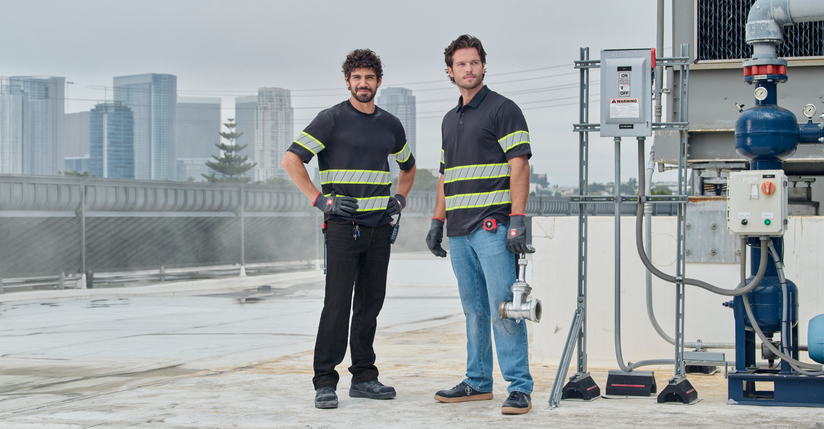Two men wearing black shirts with yellow reflective stripes stand on a rooftop near machinery, with a city skyline visible in the background. One holds a pipe fitting; both wear gloves and look at the camera.