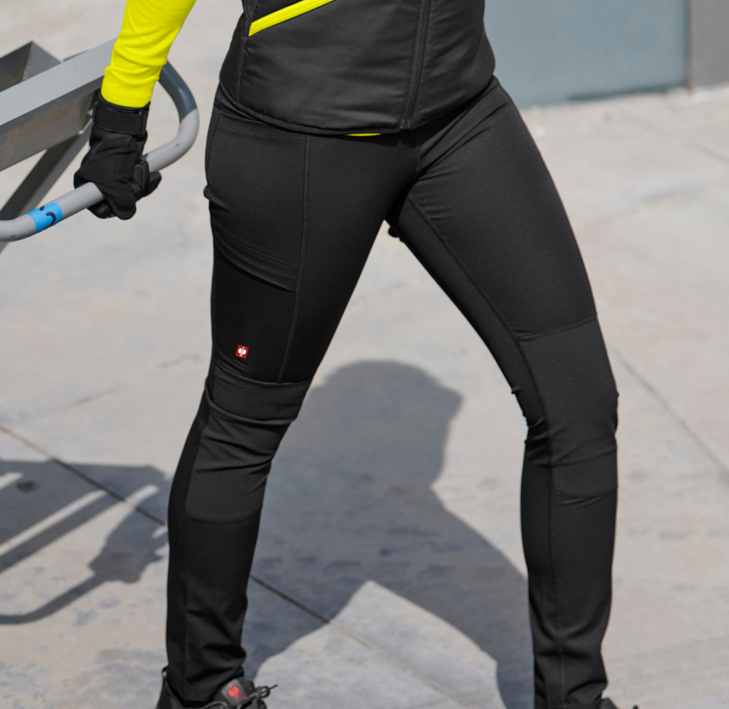 Person wearing black athletic pants and a yellow-sleeved top stands outdoors on pavement, holding onto a metal railing with gloved hands. Their face is not visible in the image.