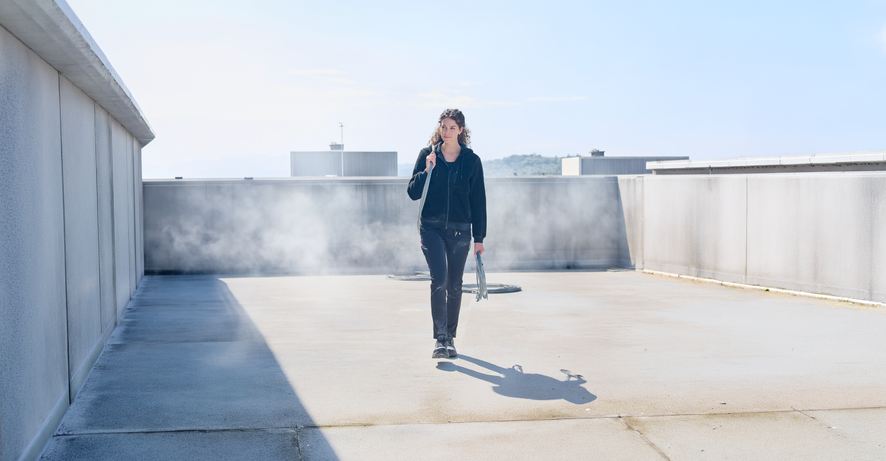 A person dressed in black walks on a rooftop under a clear sky, holding an umbrella and casting a shadow on the sunlit concrete, with mist rising in the background.