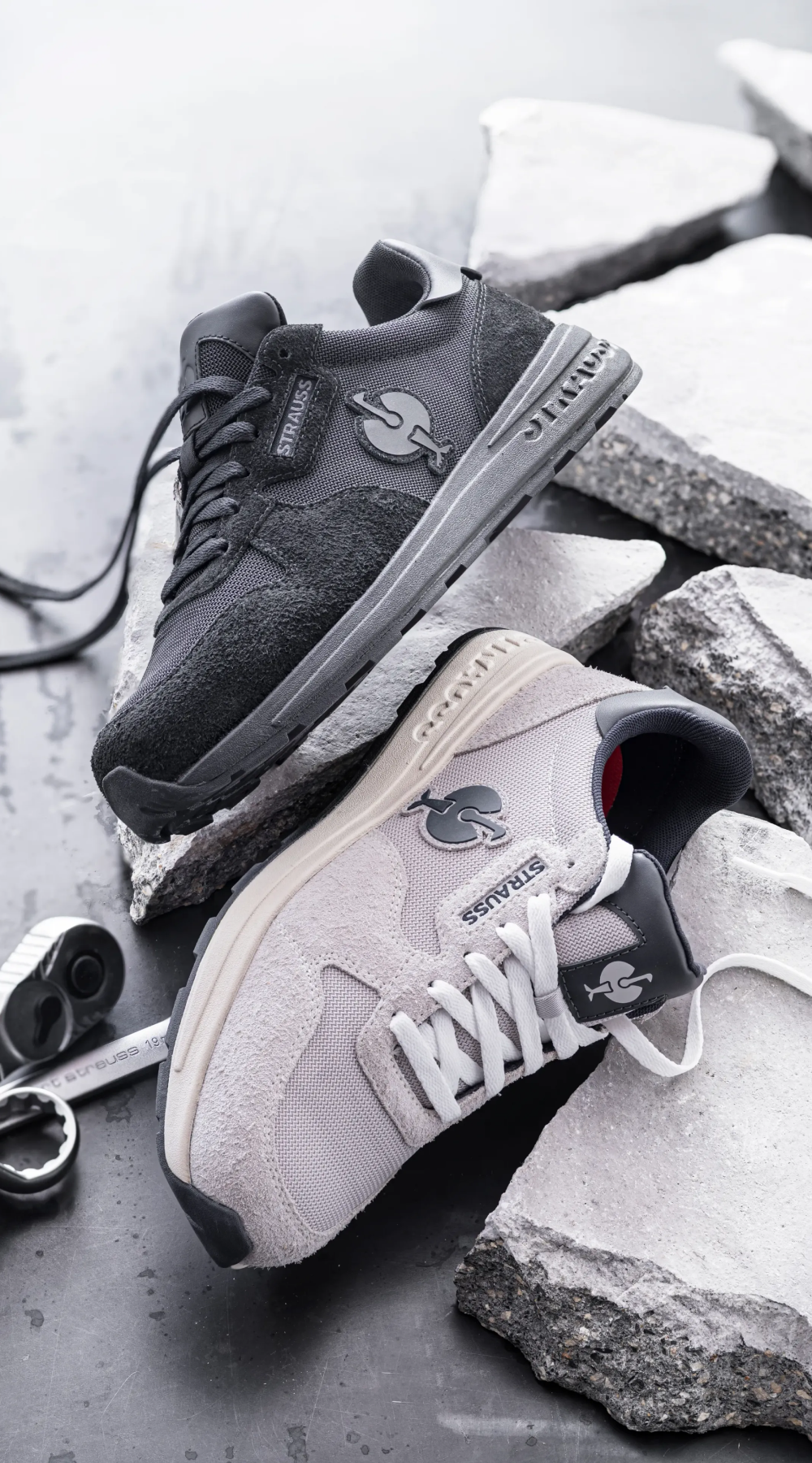 Two sneakers, one black and one light gray, rest on broken concrete slabs. Both shoes feature the Jurassic Park logo on the side. A metal tool and shoelace are also visible beside the sneakers.