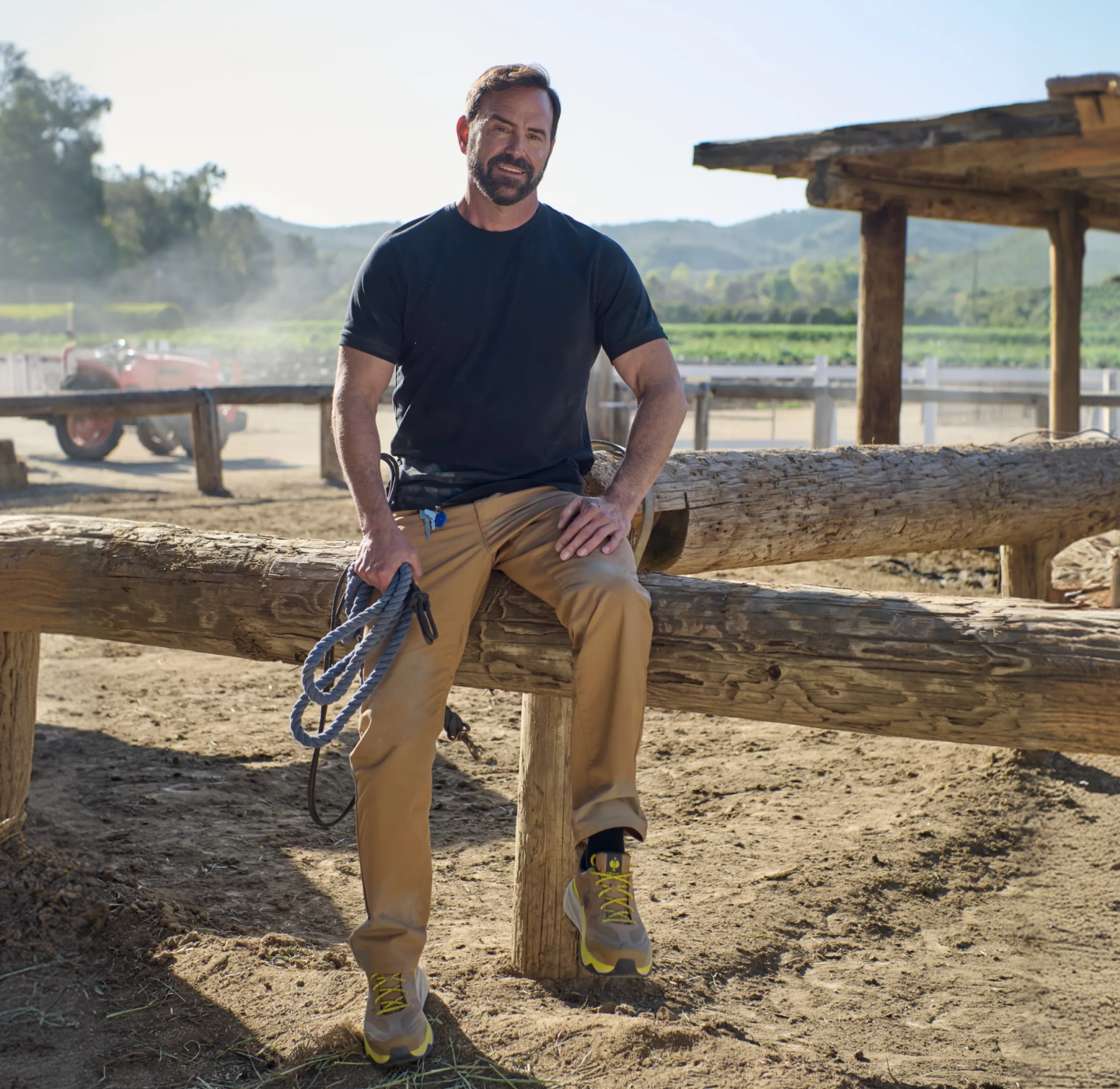 A man in a black t-shirt and tan pants sits on a wooden fence at a ranch, holding a rope. Theres a tractor, dirt ground, and hills in the background under a clear sky.