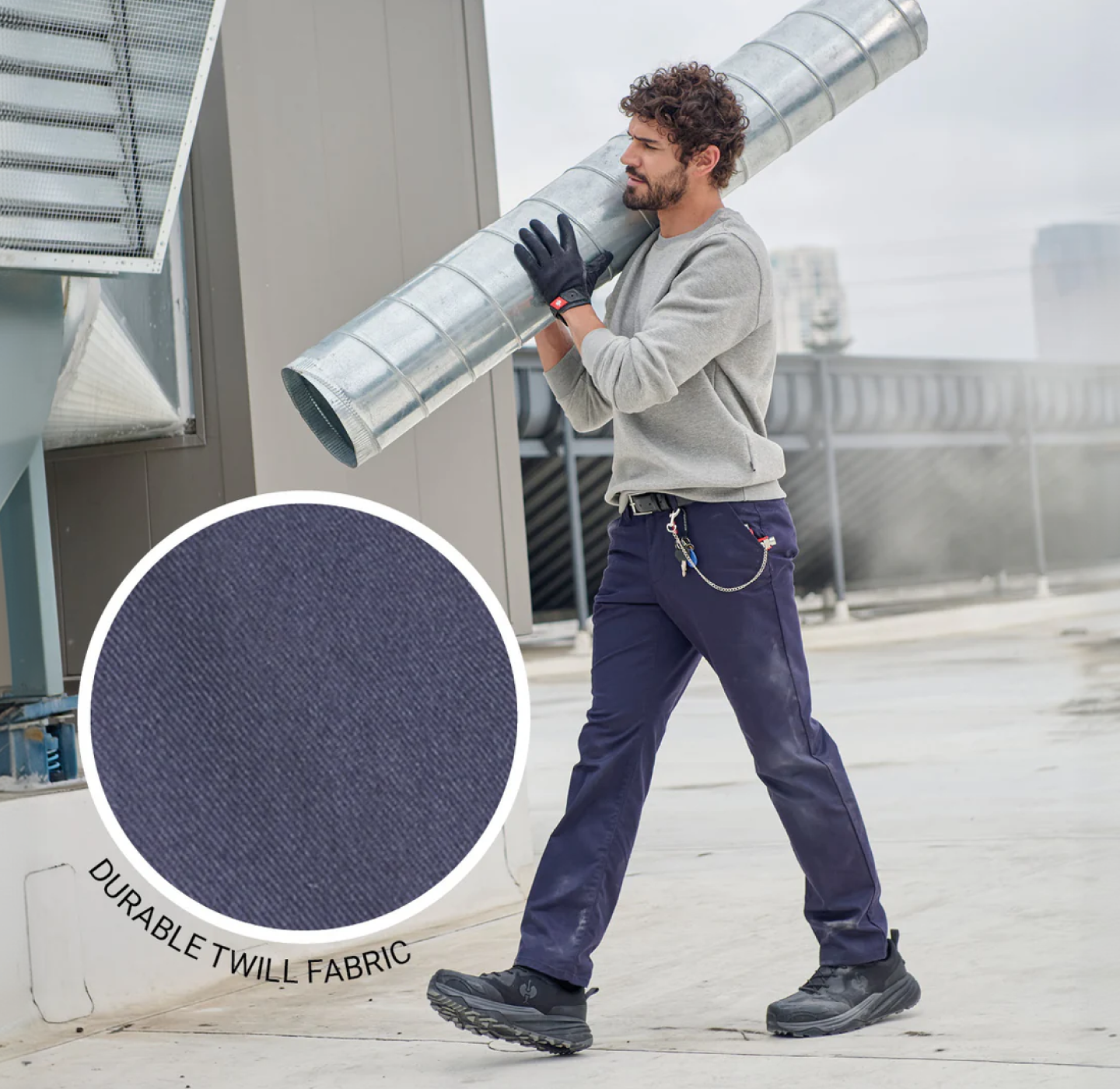 A man in work boots and navy pants carries a large metal duct on his shoulder at an industrial site. An inset shows a close-up of durable twill fabric used in the pants.