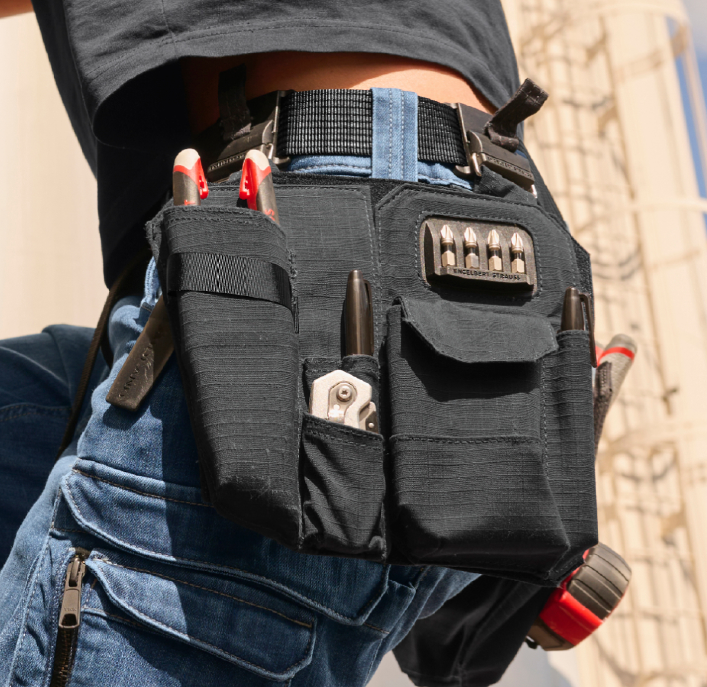 A close-up of a person wearing a tool belt with a tape measure, pliers, and screwdrivers. The person is dressed in a black shirt and dark pants.