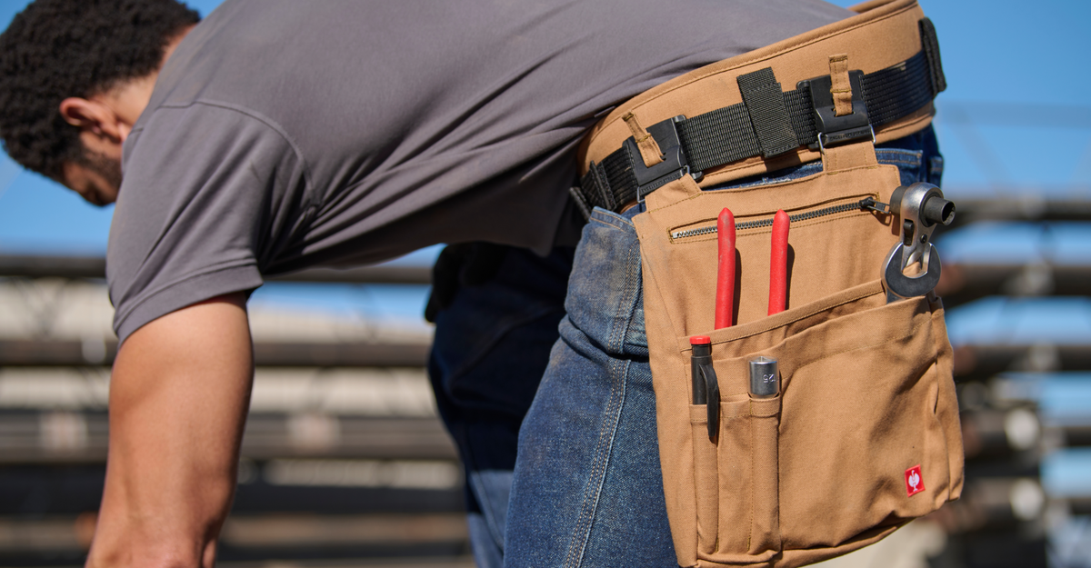 A person wearing a tool belt with pliers, a wrench, and other tools in the pockets, bending over while working outdoors.