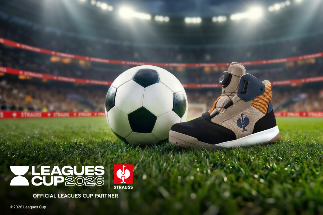 A beige, black, and orange safety work boot stands on a green soccer field next to a classic black-and-white soccer ball, with a blurred stadium in the background. Leagues Cup 2026 and Strauss logos are visible in the lower-left corner.