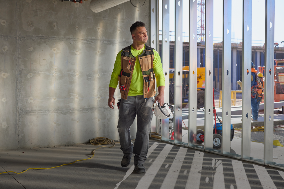Man walking in a construction site wearing a high-visibility vest with reflective stripes.