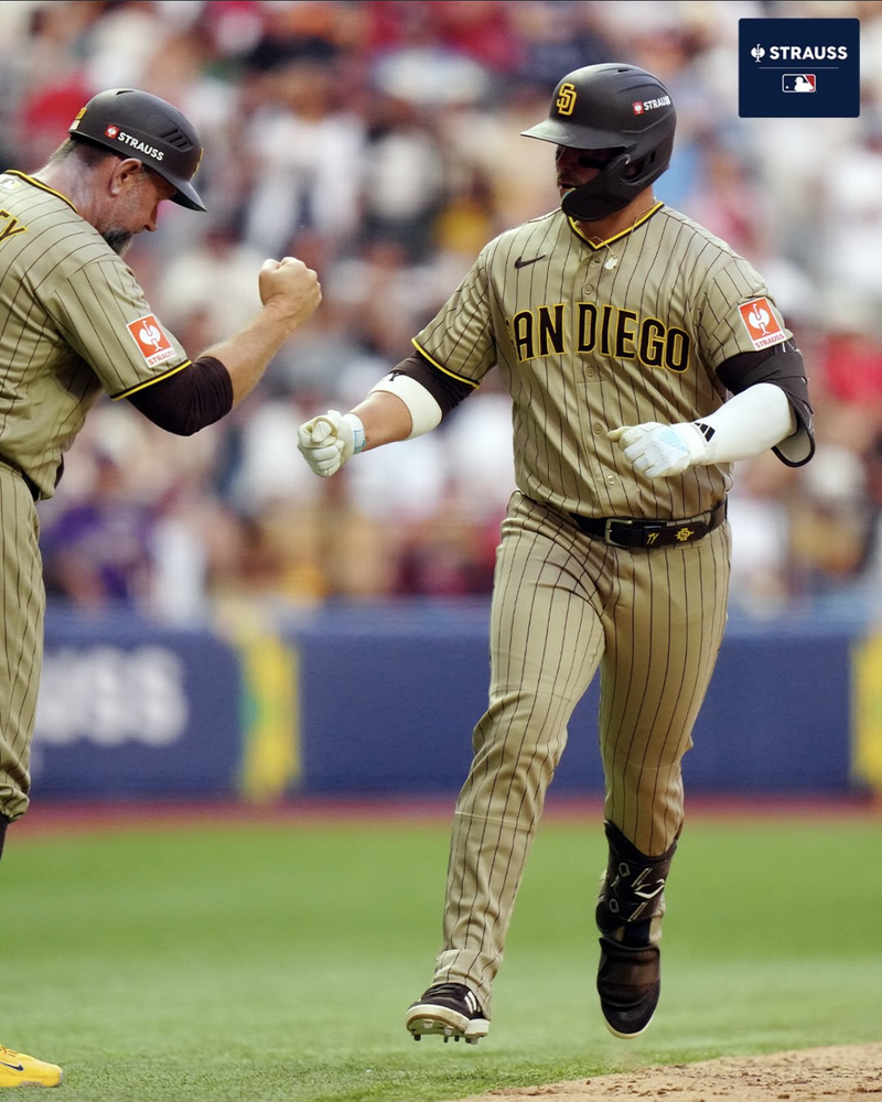 Two padres players bump fists as one rounds the base at the CDMX series in 2026. A Strauss logo is on the sleeve and helmet of the players. 