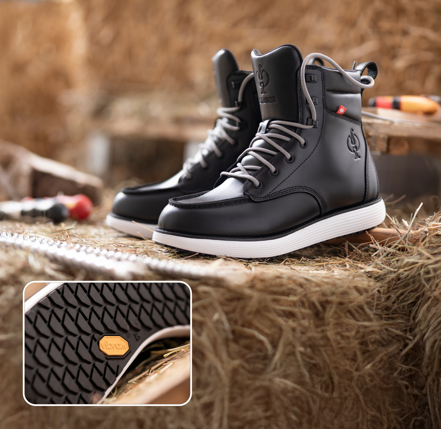 A pair of black boots with white soles displayed on hay, with a close-up inset showing the sole pattern and a small yellow logo. Tools and straw are visible in the blurred background.