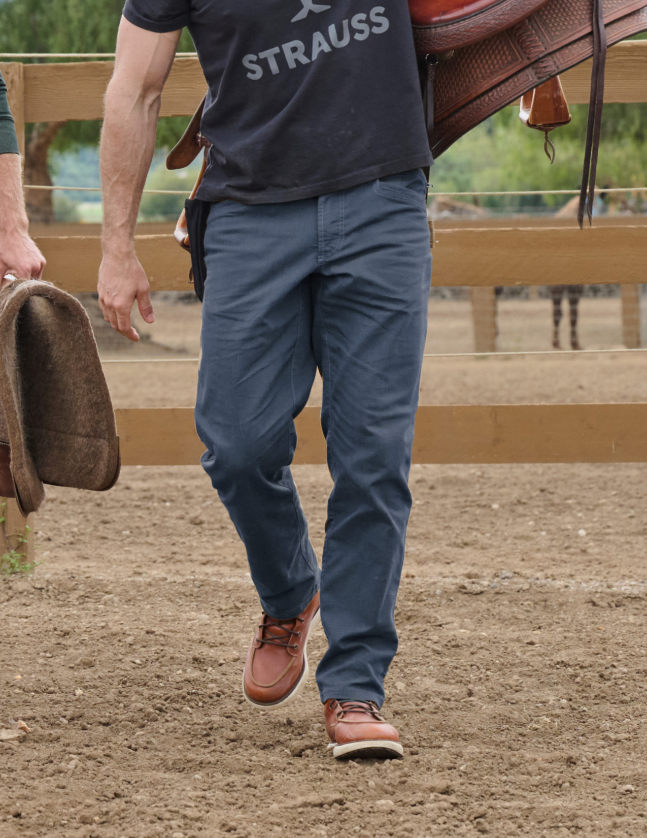 Person wearing a navy shirt with 'STRAUSS' branding, blue pants, and brown shoes walking on a dirt path.