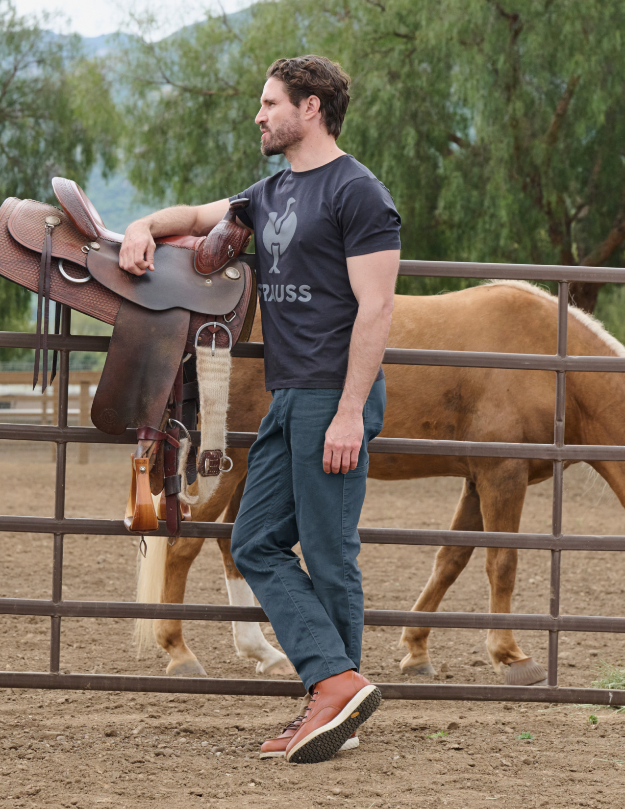 Man holding a horse saddle in front of a horse in an outdoor setting
