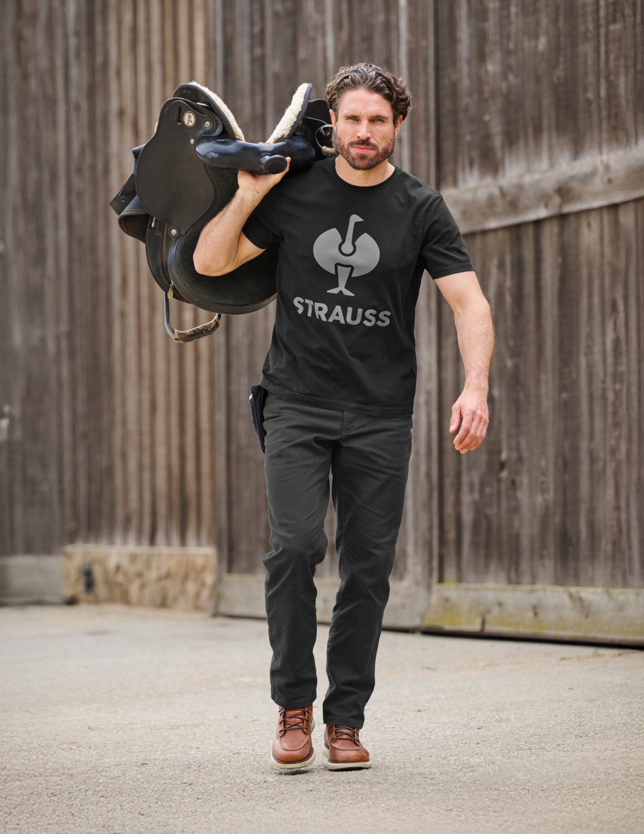 Man carrying a horse saddle while wearing a black t-shirt with 'Strauss' logo and black pants.