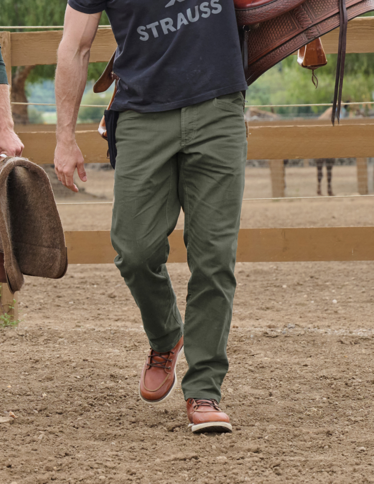 Person wearing a navy shirt with 'Strauss' branding, green pants, and brown shoes walking on a dirt path with a horse in the background.