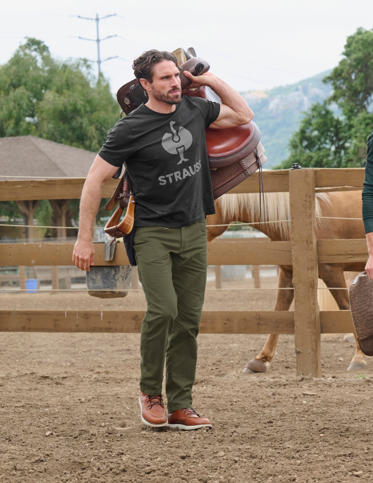 Man wearing green pants holding a saddle standing in an outdoor setting with horses and a wooden fence.