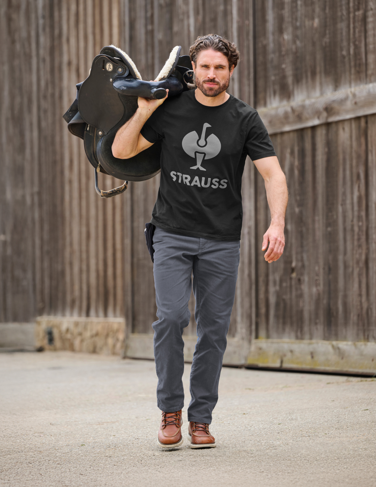 Man carrying a horse saddle while wearing a black t-shirt with 'Strauss' logo and grey pants.