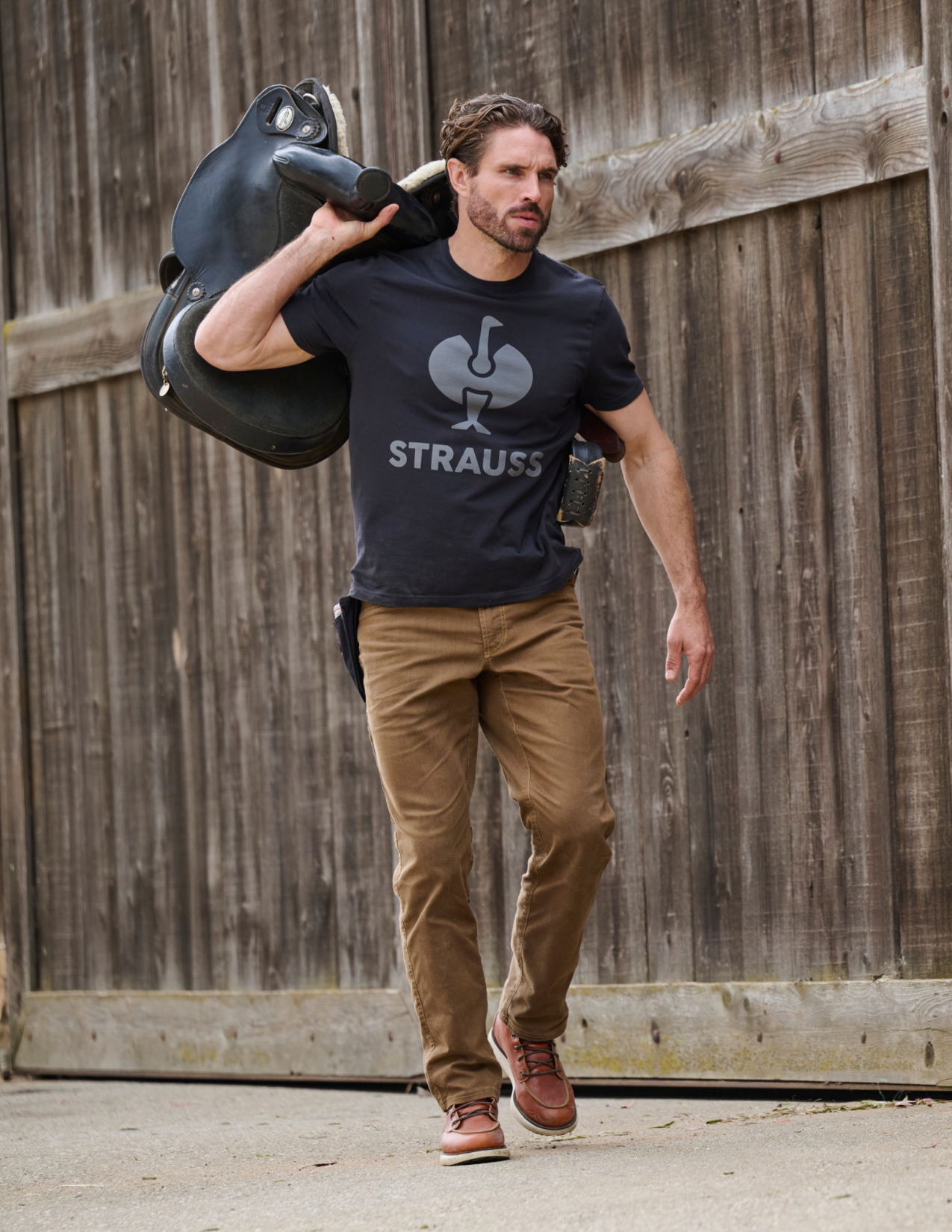 Man carrying a large black object, wearing a black t-shirt with 'STRAUSS' logo and brown pants, against a wooden fence background.